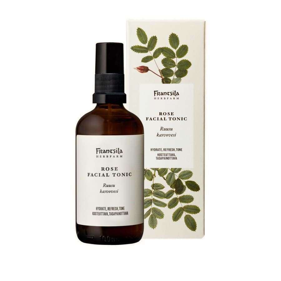Frantsila Ruusu Kasvovesi; ruskea suihkepullo tekstillä "Frantsila Herb Farm Rose Facial Tonic" seisoo pakkauksensa edessä, molemmissa sama etiketti ja kasvikuvitus ruusunvarresta lehtineen ja nuppuineen Frantsila Herb Farmilta; pullossa musta muovinen suihkekorkki; sekä pullo että rasia mattapintaisia; etiketeissä serif-fontti; neutraali tausta, ei ihmisiä.