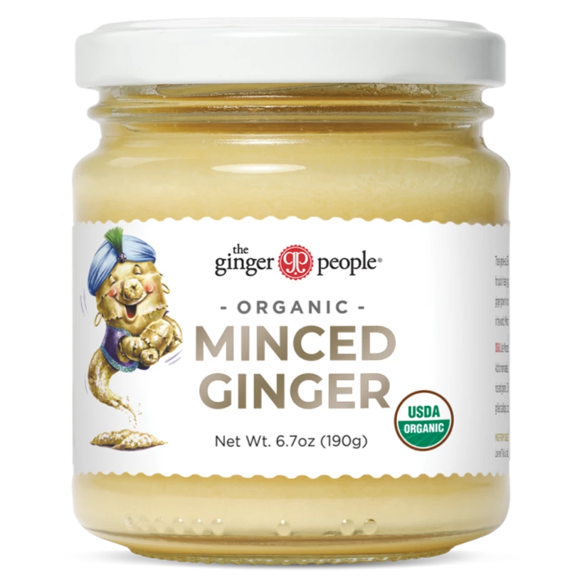 The Ginger People luomu murskattu inkivääri; kirkas lasipurkki metallikannella, valkoisessa etiketissä sarjakuvamainen inkiväärihahmo ja USDA Organic -sertifiointimerkki; lihavoitu, sekakirjaiminen groteskiteksti tuotenimestä; valkoinen tausta.
