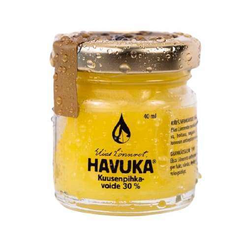 HAVUKA Kuusenpihkavoide 30% -lasipurkki, sisältää keltaista voidetta; kirkas lasi, sylinterimäinen muoto, näkyvissä kondenssikosteutta ulkopinnalla; kansi metallisen kultainen ja pyöreä, kiinnitetty kultaisella tarralla kannen yli; etikettiteksti yksinkertaista ja suuraakkosin; ei ihmisiä tai käsiä näkyvissä; tausta yksinkertainen.