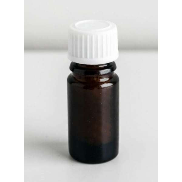 Helios Arnica Montana -pullo; pieni meripihkanvärinen lasipullo valkoisella muovisella kierrekorkilla, pystyssä valkoisella alustalla; pullo sylinterimäinen sileällä ja kiiltävällä pinnalla; ei näkyvää etikettiä tai tekstiä; kuva otettu silmän tasolta tasaisessa valaistuksessa yksivärisellä valkoisella taustalla; ei ihmisiä tai käsiä näkyvissä.