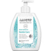 Lavera Naturkosmetik Gentle Care Mild Hand Wash -käsipesu; valkoinen ja sininen pumppupullo vesiroiskekuvituksella ja tuotetiedoilla etiketissä; lieriömäinen muovipullo kiiltävällä pinnalla, vaaleansininen pumppu; etiketissä "Lavera Naturkosmetik" lihavoidulla groteskifontilla; ei ihmisiä tai käsiä; vaalea tausta, suoraan edestä kuvattu studiokuva.