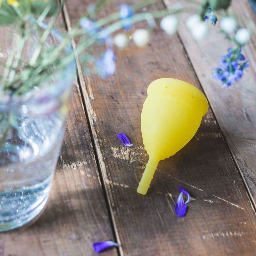 Lunette Kuukuppi Keltainen puisella alustalla, vieressä lasi vettä ja violetteja kukkia sekä hajallaan olevia terälehtiä