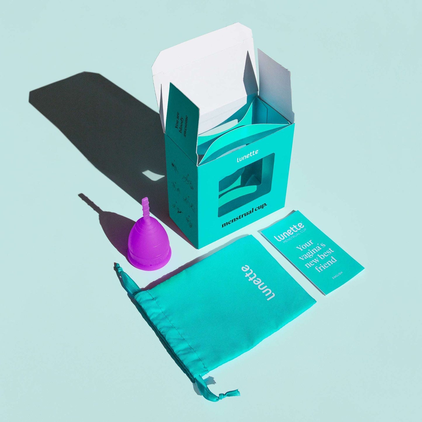 Violetti Lunette-kuukuppi, teal-värinen kangaspussi, pieni valkoinen ohjevihko ja avattu teal-värinen "Lunette"-pahvirasia aseteltuna vaalean sinisen pinnan päälle