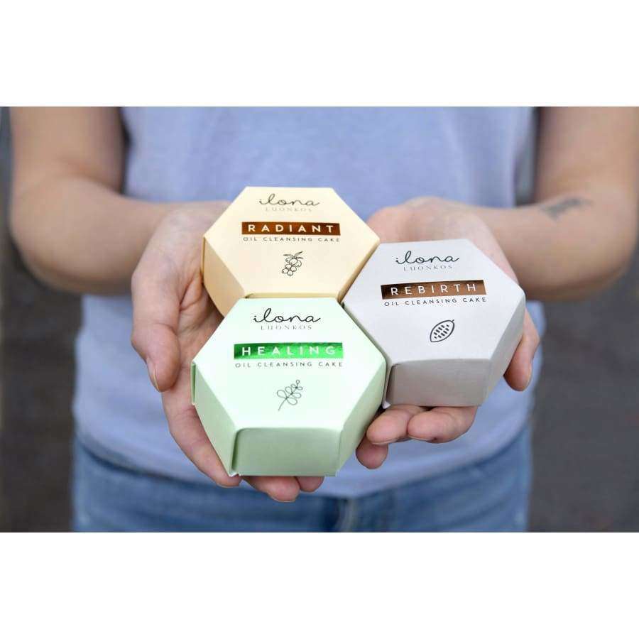 Henkilö pitelee kolmea kuusikulmaista laatikkoa nimeltä Radiant, Healing ja Rebirth; pastellinkeltaiset, -vihreät ja beiget laatikot esittelevät kukin Ilona Lux Studio Oil Cleansing Cake -tuotteen etupuolellaan; jokainen laatikko mattapintainen minimalistisin tekstausvapain valkoisin etiketein; ympäristö vaalea, pehmeästi luonnonvalossa valaistu; laatikot pinottuna yhteen käteen täyttäen suurimman osan kuvasta.