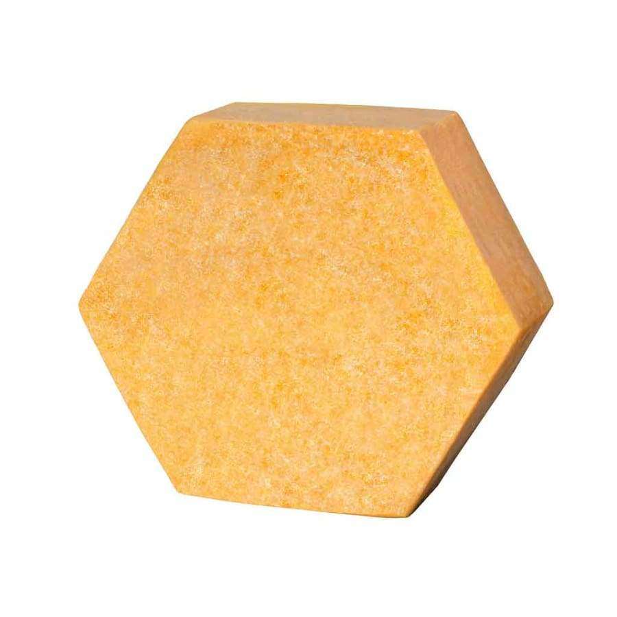 Luonkos Celebrate Sparkling Body Cake; kiinteä, kuusikulmainen keltaoranssi kappale karkealla pinnalla, muistuttaen katukiveä, eristettynä valkoiselle taustalle; ei näkyvää etikettiä tai pakkausta; pinta matta ja karkea; ei tekstiä, brändimerkintöjä, ihmisiä tai toimintaa; studiokuva tasaisella valaistuksella korostaa esineen muotoa ja pintaa.