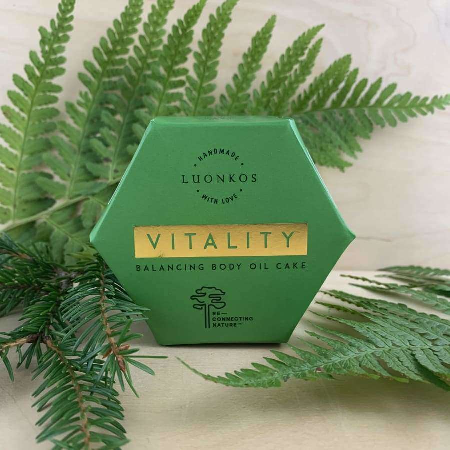 Luonkos VITALITY Vartaloöljykakku; vihreä kuusikulmainen rasia tekstillä "LUONKOS VITALITY Balancing Body Oil Cake" mattapahvilla, sijoitettuna vaalealle puupinnalle; typografia lihavoitu ja groteski, valkoinen väri; useita vihreitä saniaisenlehtiä kehystää rasiaa; ei ihmisiä tai käsiä; asetelmakuva puisella alustalla, ympärillä tuoreita saniaisenlehtiä; öljykakku tuo luonnollista elinvoimaa ihonhoitorutiiniin.