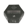 Luonkos Complete Facial Oil Cleansing Cake For Men; kuusikulmainen musta rasia, jossa yksinkertaisia kasvojenpuhdistuskuvituksia kyljessä; rasia mattapintaista pahvia, kauttaaltaan mustaa pintaa, valkoista keskitettyä groteskitekstiä etusivulla; puhtaan geometrinen pakkaus kuvattu vaalealla taustalla, ei ihmisiä tai rekvisiittaa.