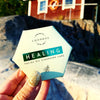 Käsi kiiltävällä kultaisella kynsilakalla pitelee kuusikulmaista LUONKOS HEALING Facial Oil Cleansing Cake -pakkausta ulkona, taustalla kiviä ja punainen puurakennus; pakkaus mattapintainen, vaaleasävyinen mustalla tekstausvapaalla tekstillä; kuva otettu ulkona luonnonvalossa, taustalla näkyy kiviä ja puuverhoilua.
