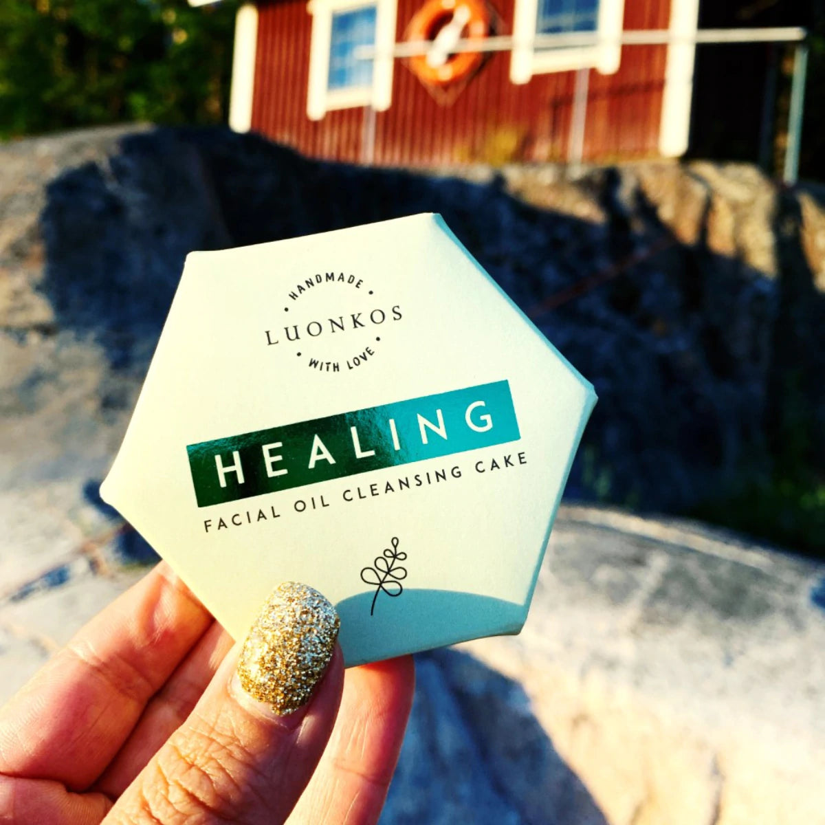 Käsi kiiltävällä kultaisella kynsilakalla pitelee kuusikulmaista LUONKOS HEALING Facial Oil Cleansing Cake -pakkausta ulkona, taustalla kiviä ja punainen puurakennus; pakkaus mattapintainen, vaaleasävyinen mustalla tekstausvapaalla tekstillä; kuva otettu ulkona luonnonvalossa, taustalla näkyy kiviä ja puuverhoilua.