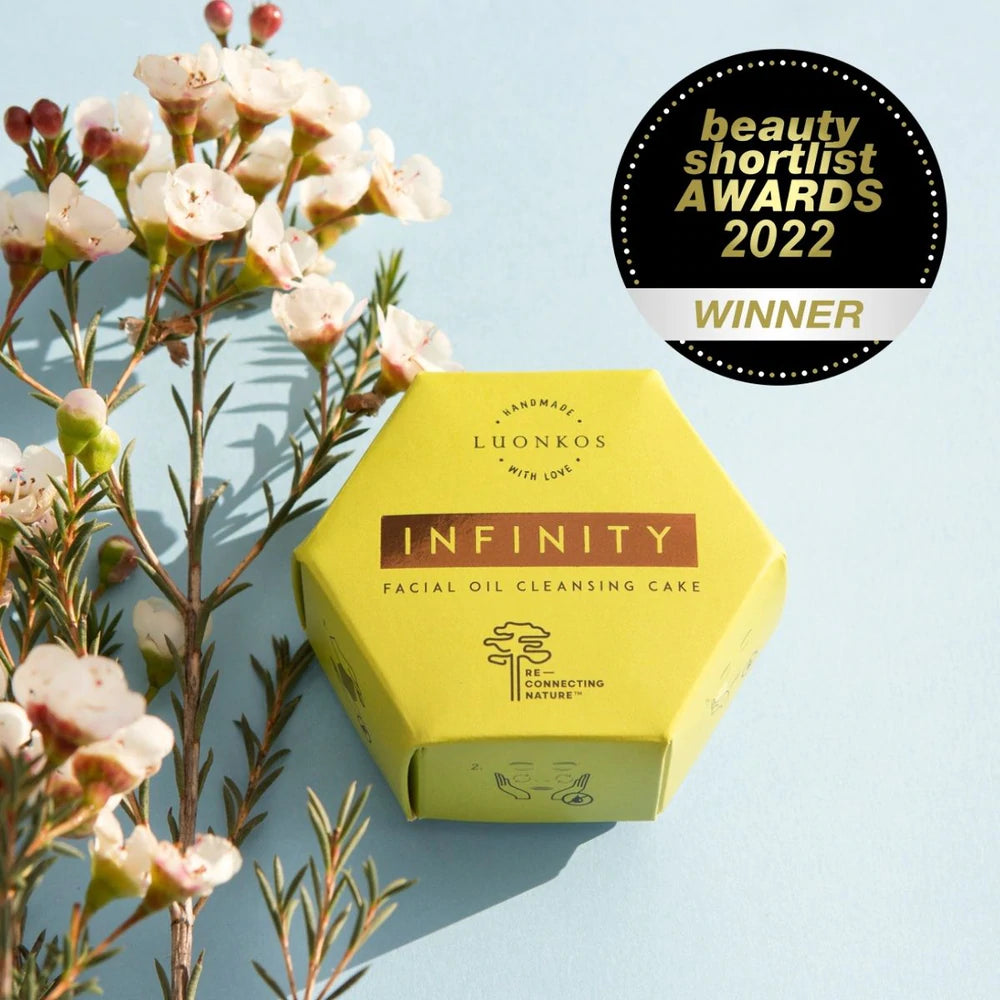 Luonkos INFINITY Öljypuhdistuskakku; keltainen kuusikulmainen rasia tekstillä "Infinity Facial Oil Cleansing Cake" by Luonkos valkoisten kukkien vieressä; pyöreässä merkissä lukee "beauty shortlist AWARDS 2022 WINNER"; mattapintainen pakkaus, suuri groteskiteksti mustana; kuva otettu suoraan edestä sinistä taustaa vasten, tuote keskitettynä, ei ihmisiä tai käsiä; tuote erottuu selvästi sinisestä taustasta.