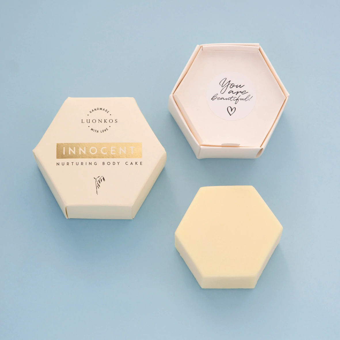 Luonkos Innocent Nurturing Body Cake; kuusikulmainen kappale kermanvärisessä pakkauksessa, avatun kannen sisäpuolella teksti "You are beautiful" sydämen kera; tuote ja pakkaus aseteltuna rinnakkain vaaleansinisellä alustalla; molemmissa mattapinta ja selkeä groteskiteksti; ei ihmisiä tai käsiä kuvassa.