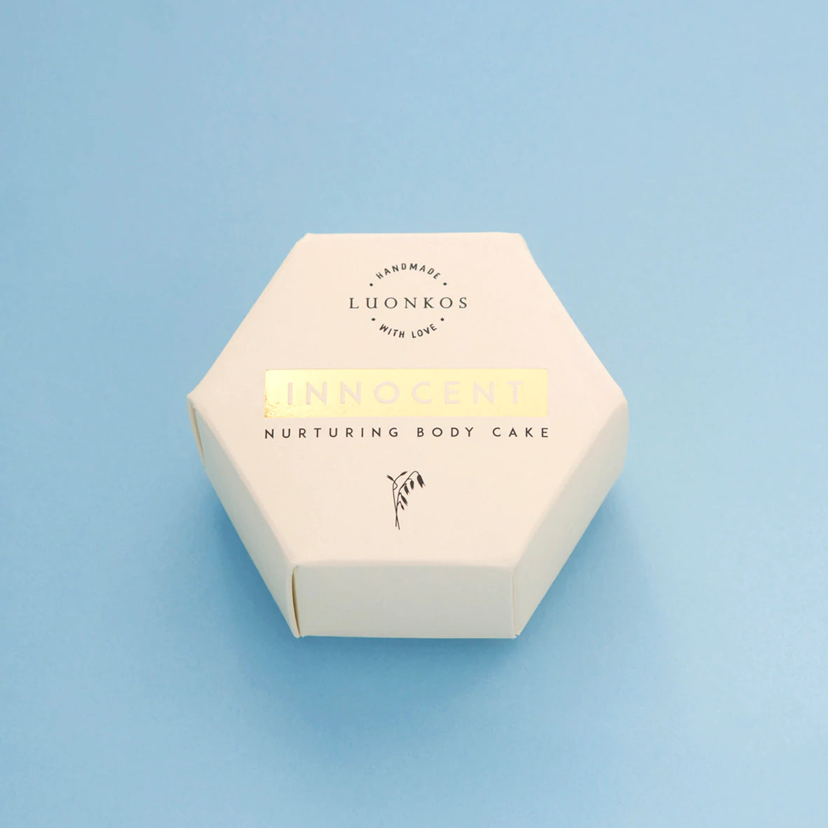Luonkos Innocent Nurturing Body Cake; kuusikulmainen valkoinen rasia vaaleansinisellä taustalla; rasia mattavalkoinen; teksti minimalistinen, mustaa kultaisin korostuksin, keskitettynä etupaneeliin; ei ihmisiä tai käsiä; kuva otettu suoraan ylhäältä näyttäen vain suljetun pakkauksen ja taustan.