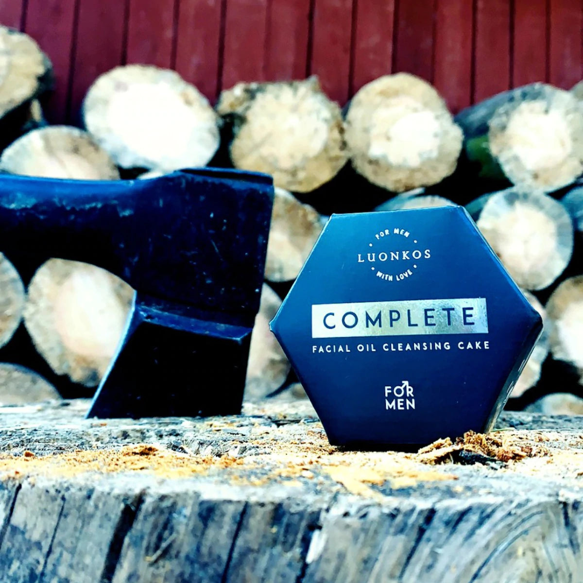 Luonkos Complete Facial Oil Cleansing Cake For Men; kuusikulmainen kasvojenpuhdistuspala pystyssä karkeasti sahatun puun päällä, vieressä puukahvainen kirves; tuote kermanvärinen, matta pinta, selkeää lihavoitua groteskitekstiä; taustalla pinottuja halkoja ja punaiseksi maalattu seinä; ei ihmisiä tai käsiä näkyvissä.