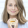 Olkapään mittaisia vaaleita hiuksia omaava nainen hymyilee pehmeästi pidellen pientä suorakulmaista laatikkoa nimeltä Ilona Lundenkos Rebirth Oil Cleansing Cake, esitellen hehkuvaa ja tervettä ihoa vaaleaa taustaa vasten; laatikko valkoinen ja yksinkertaisin keskitetyin tekstausvapain mustin tekstein; asetelma tasaisesti valaistu ilman näkyvää rekvisiittaa tai huonekaluja.