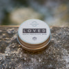 Luonkos LOVED Valkosuklaabalsami; pyöreä metallinen purkki mattapintaisena lumisen puisen pinnan päällä, tuoksuna valkosuklaa; etiketissä selkeä versaalikirjaimin painettu teksti; ei ihmisiä tai käsiä kuvassa; ulkotilassa kuvattu tausta, jossa näkyy lunta karkealla puulaudoituksella.