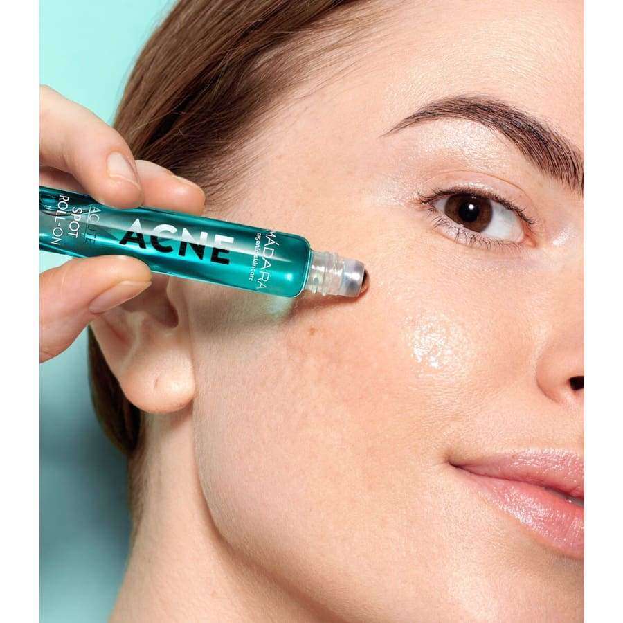 Mádara ACNE Acute Spot Roll-On; henkilö levittää sinistä täsmähoitopuikkoa poskeensa, paljastaen sileän, kiiltävän ihon lähikuvassa vaaleansinistä taustaa vasten; lieriömäinen roll-on-tuote, kiiltävä sininen muovipakkaus, valkoinen korkki ja painojälki; käsi pitelee levitintä; ei muita esineitä näkyvissä; etikettitekstiä minimaalisen luettavissa groteskifontilla.