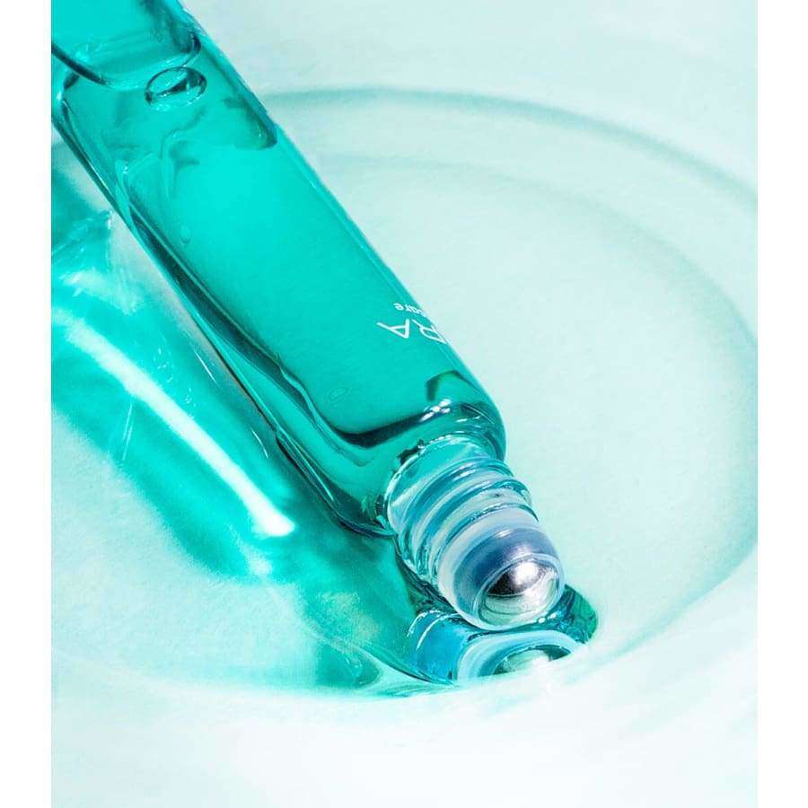 MÁDARA ACNE Acute Spot Täsmähoito Roll-on; lähikuva turkoosista lasisesta rullapullosta vaaleansinisellä pinnalla, korkki irti ja metallinen rulla paljastuneena, valoa heijastaen; pullo lieriömäinen, kiiltäväpintainen; ei näkyvää tekstiä tai etikettejä tuotteessa; kuvassa vain pullo, ei ihmisiä, käsiä tai toimintaa; tausta tasainen ja sileä.