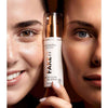 MÁDARA FAKE IT Healthy Glow Self Tan Serum; kaksi naista pitelee pulloa kasvojensa välissä, korostaen erilaisia ihonsävyjä ja säteilevää, tervettä ihoa luonnollisella meikillä; pullo lieriömäinen, muovia, satiininen pinta ja pumppuannostelija; etiketissä lihavoitua suuraakkosista groteskitekstiä tuotenimelle; neutraali tausta; lähikuva keskittyy kasvoihin ja käsiin.