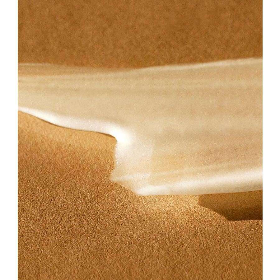MÁDARA FAKE IT Healthy Glow Self Tan Serum; lähikuva kermaisesta, valkoisesta emulsiosta levitettynä tekstuuriselle, vaaleanruskealle pinnalle; emulsion koostumus sileä ja hieman kiiltävä; ei näkyvää pakkausta tai luettavaa tekstiä; ei ihmisiä tai käsiä; kuva keskittyy tiiviisti tuotteeseen ja taustan tekstuuriin.