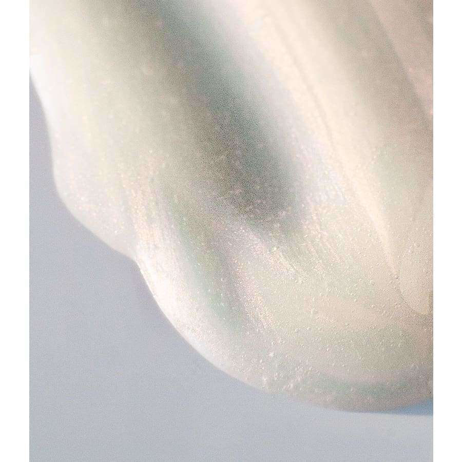 Mádara Melting Cleansing Oil -puhdistusöljy; lähikuva sileästä, helmiäisenvalkoisesta pinnasta hienovaraisin kiiltävin korostuksin ja pehmein kaarevin reunoin vaaleaa taustaa vasten; esineessä kiiltävä pinta ja pyöreä muoto; ei näkyvää pakkausta, etikettitekstiä tai ihmisiä.