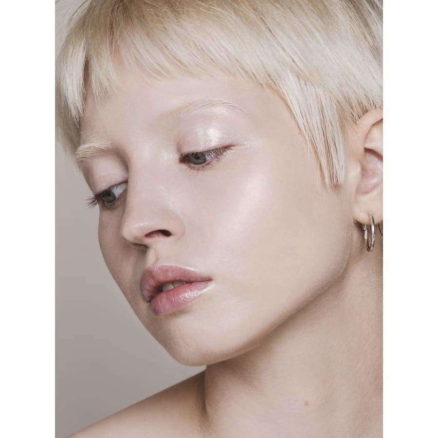 MÁDARA Skin Equal Meikkivoide, sävy Porcelain; henkilö, jolla lyhyet vaaleat hiukset, kevyt meikki ja kimmeltävä iho, katsoo alaspäin; pieni rengaskorvakoru; pehmeä, neutraali ilme vaalealla taustalla.