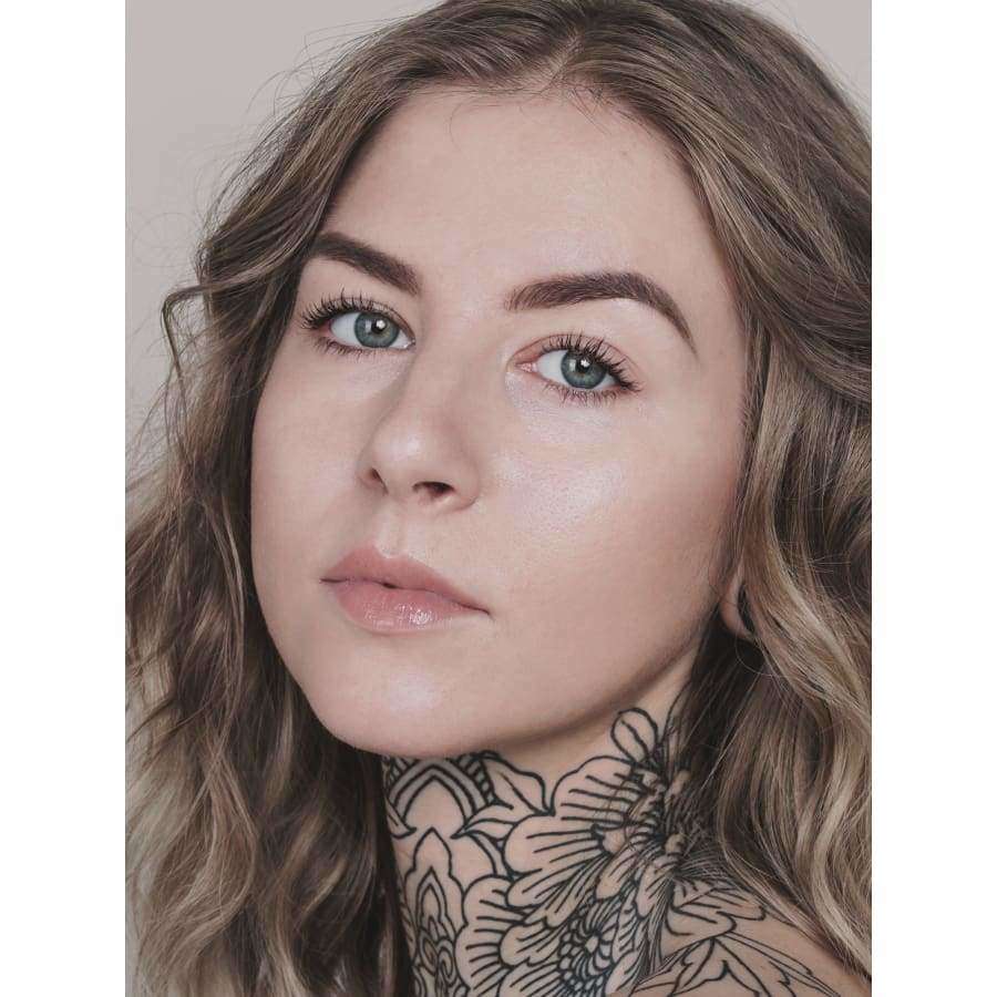 MÁDARA Skin Equal Meikkivoide, sävy Rose Ivory; nuori nainen, jolla aaltoilevat vaaleanruskeat hiukset ja siniset silmät, katsoo kameraan; luonnollinen meikki sekä suuri kukkatatuointi kaulassa; vaalea, neutraali tausta; ei näkyvää pakkausta tai tekstiä.