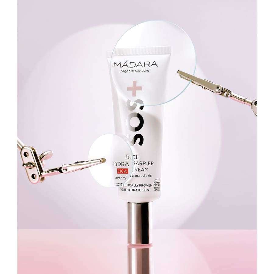 Madara SOS Rich Hydra-Barrier CICA Kasvovoide; MÁDARA SOS Rich Hydra-Barrier CICA Cream -tuubi pystyssä, kiinnitettynä metallipuristimilla; tuubi mattavalkoinen pystysuoralla vaaleanpunaisella raidalla ja mustalla groteskitekstillä; korkki kiiltävän valkoinen; kaksi lasista ympyrää suurentaa etikettitietoja; tausta pehmeän vaaleanpunaisen ja violetin sävyinen; ei ihmisiä tai käsiä kuvassa.