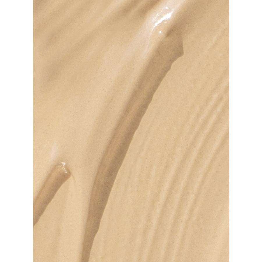 MÁDARA The Concealer, sävy Latte; lähikuva sileästä, beigenvärisestä BB-voiteesta tai nestemäisestä meikkivoiteesta, näkyvissä sivellinjälkiä ja kiiltävä pinta; kermainen koostumus, vaalea beige väri; ei pakkausta tai etikettitekstiä; vaalea tausta, ei ihmisiä tai käsiä; tiivis lähikuva.