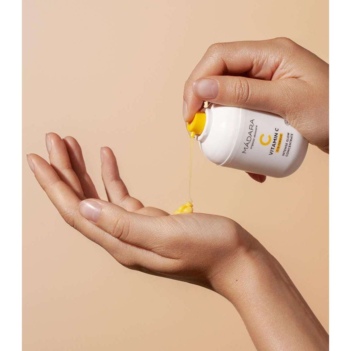 Madara Vitamin C Heleyttävä C-vitamiinikonsentraatti; henkilö annostelee keltaista seerumia valkoisesta pullosta tekstillä "MÁDARA Vitamin C concentrate" avoimeen kämmeneen beigellä taustalla; pullo sylinterinmuotoinen mattapintainen mustalla groteskitekstillä; korkki valkoista muovia; minimalistinen asetelma, lähikuva kädestä, tuotteesta ja nesteestä.