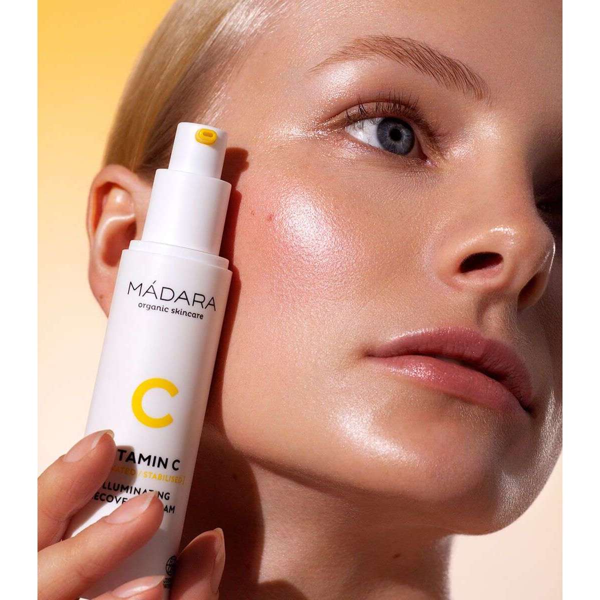 Madara Vitamin C Kirkastava C-vitamiinivoide; henkilö, jolla hehkuva ja säteilevä iho, pitelee MÁDARA Vitamin C Illuminating Recovery Cream -pulloa lähellä kasvojaan; pullo lieriömäinen, valkoinen, pumppuannostelijalla ja mattapinnalla; tuotenimi painettu selkeällä groteskifontilla etiketissä; tausta pehmeän keltaisesta valkoiseen liukuvärinen; lähikuva silmän tasolta.