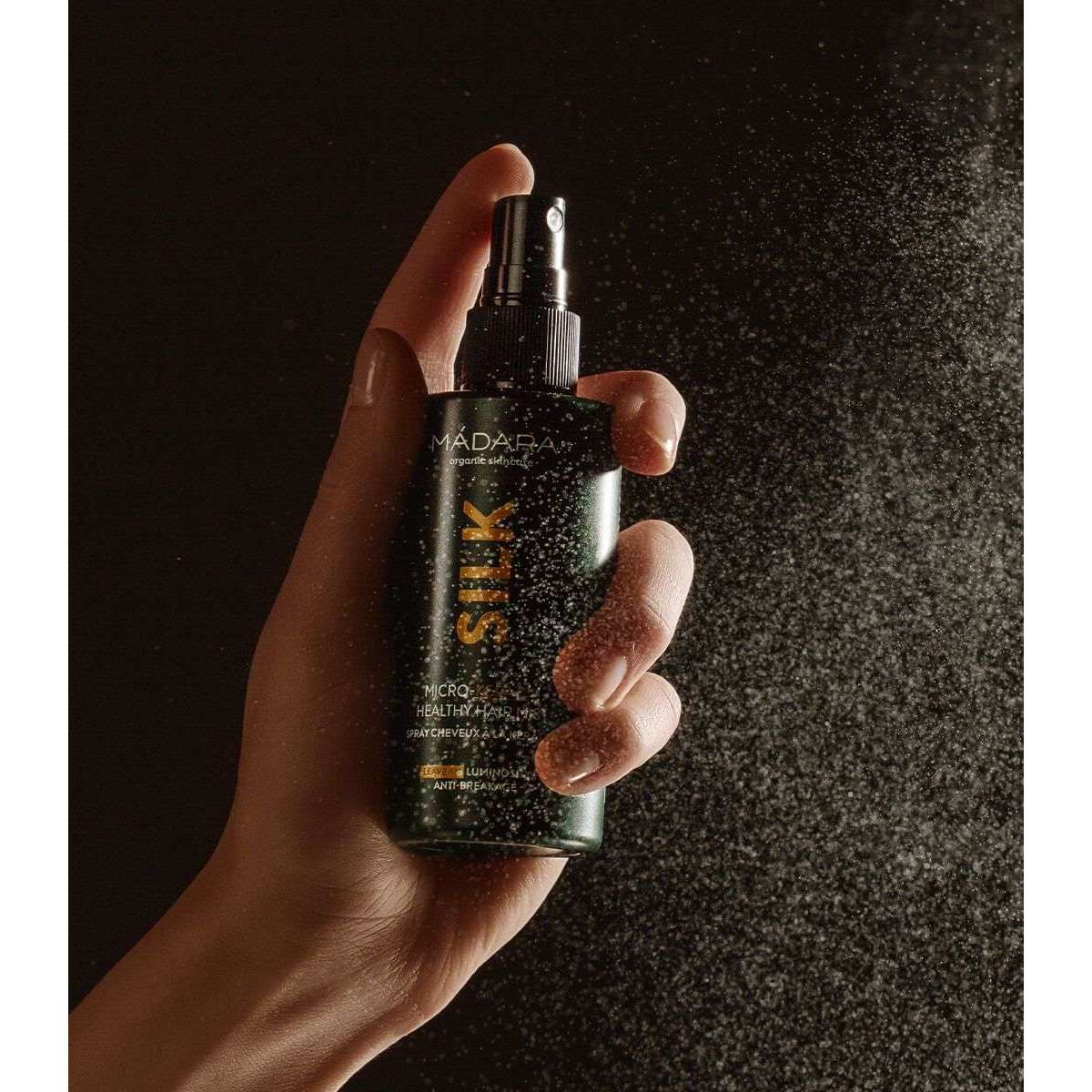 Madara SILK Micro-Keratin Keratiinisuihke; käsi pitelee tummaa mattapintaista sylinterinmuotoista suihkepulloa; etiketissä teksti "SILK" lihavoidulla groteskikirjasimella; suuttimesta suihkuaa hieno sumu valon läpäisemänä; tausta täysin musta, kuvassa näkyy vain käsi ja osa käsivarrestä lähikuvana tuotteesta ja suihkeesta.