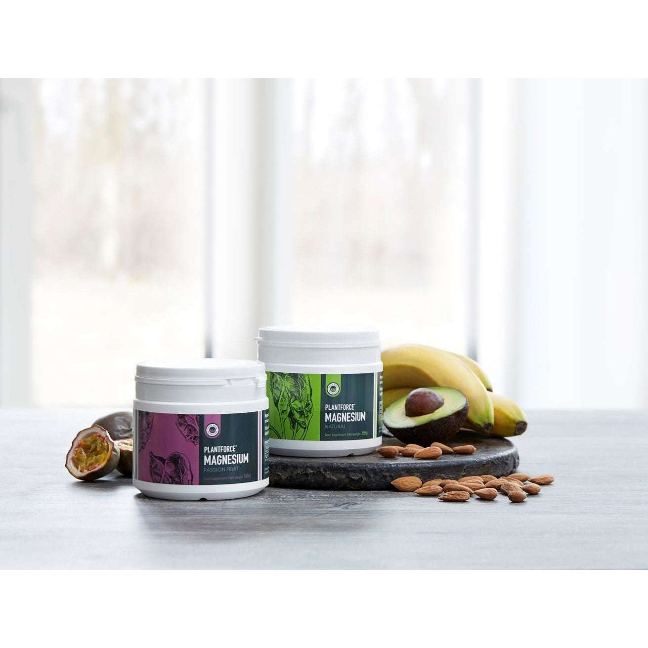 Plantforce Magnesium Maustamaton; kaksi valkoista sylinterinmuotoista purkkia tekstillä "Magnesium" selkeällä groteskikirjasimella, aseteltuna tummanharmaalle liuskekivialustalle; vierellä raakoja manteleita, halkaistu avokado siemenineen, kuorimaton banaani ja kokonainen passionhedelmä; luonnonvalo ikkunasta taustalla; ei ihmisiä tai käsiä kuvassa.
