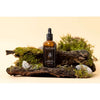 Moi Forest Dust Microbiome Magic Oil; ruskea lasinen pipettipullo tekstillä "Moi Forest Microbiome Magic Oil" aseteltuna kaarnanpalalle sammaleen, pienten kivien ja metsäaiheisen rekvisiitan kera beigeä taustaa vasten; pullo kiiltäväpinnalla ja sylinterinmuotoinen mustalla pipettikorkilla; etiketissä yksinkertaista groteskikirjasinta; lähikuva tuotteesta, ei ihmisiä tai toimintaa kuvassa.