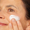 Novexpert PRO-COLLAGEN Expert Anti-Aging Cream; henkilö levittää kosteuttavaa valkoista voidetta poskelleen sormin; lähikuva keskittyy silmään, osaan nenästä ja hymyyn, iho sileän ja tasaisen näköinen; ei näkyvää pakkausta tai tuote-etikettiä kuvassa; tausta rajattu pois.