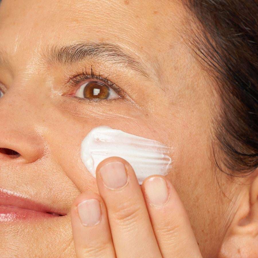 Novexpert PRO-COLLAGEN Expert Anti-Aging Cream; henkilö levittää kosteuttavaa valkoista voidetta poskelleen sormin; lähikuva keskittyy silmään, osaan nenästä ja hymyyn, iho sileän ja tasaisen näköinen; ei näkyvää pakkausta tai tuote-etikettiä kuvassa; tausta rajattu pois.