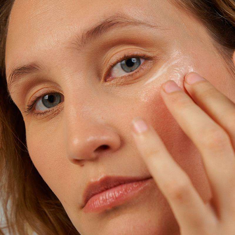 Novexpert PRO-COLLAGEN Expert Anti-Aging Eye Contour; henkilö levittää varovasti valkoista hoitovoidetta oikean silmän alle sormenpäällä; voide sileän valkoista; käsi siisti ja kynnet luonnolliset; ei näkyvää pakkausta tai etikettitekstiä; neutraali tausta, lähikuva levitysliikkeestä ja ihosta.