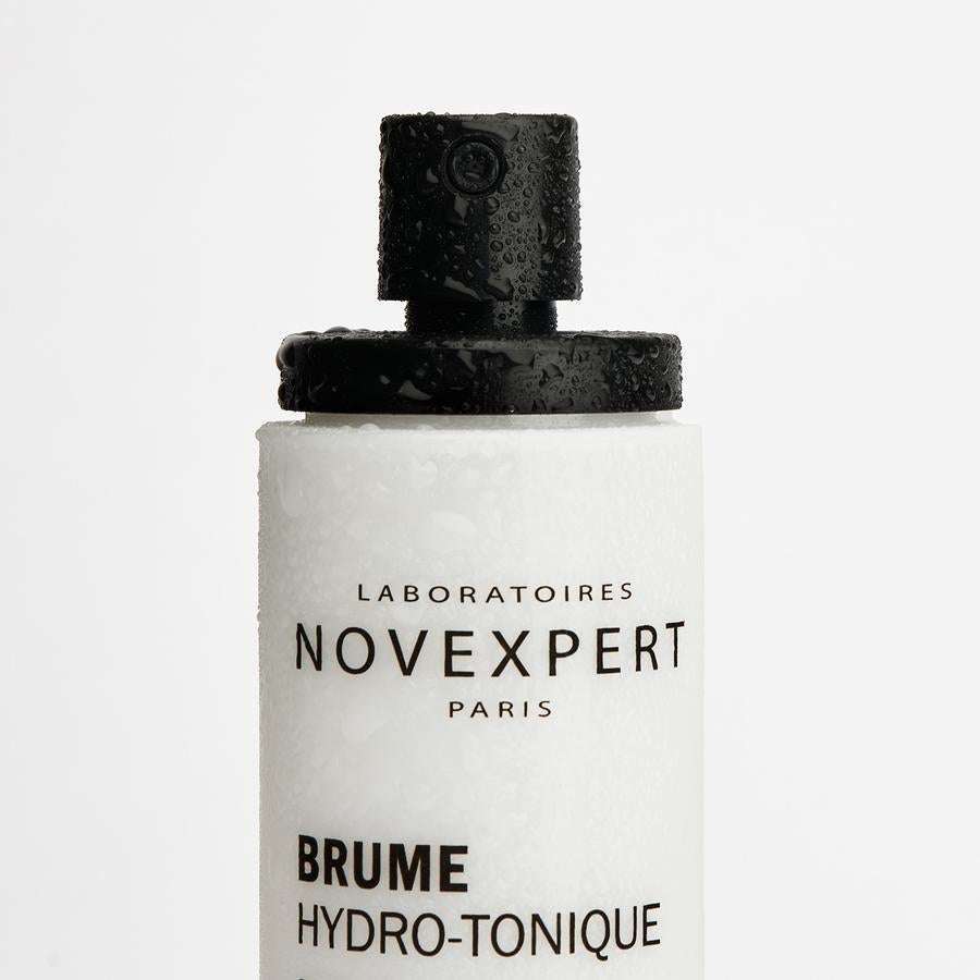 Novexpert HYALURONIC Smoothing Toning Mist; lähikuva valkoisesta muovisesta suihkupullosta tekstillä "Brume Hydro-Tonique", vesipisaroita näkyvissä mustalla mattapintaisella suuttimella ja pullon yläosassa; etiketin teksti mustalla groteskikirjasimella ja pientä oranssia yksityiskohtaa; pullo sylinterinmuotoinen; valkoinen tausta, ei ihmisiä tai käsiä.