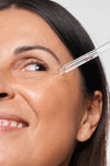 Novexpert HYALURONIC Booster Hyaluronihapposeerumi; hymyilevä henkilö levittää kosteuttavaa seerumia silmän lähelle lasisella tiputtimella katsoen ylöspäin; iho sileän ja terveen näköinen; tiputin läpinäkyvää lasia hopeisella tai metallisella korkilla; ei näkyvää etikettiä tai tekstiä; tausta neutraali, pehmeä valaistus lähikuvassa.