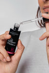 Novexpert HYALURONIC Booster Hyaluronihapposeerumi; henkilö pitelee pientä lasista tiputinpulloa ja pipettiä valmistautuen levittämään kirkasta hyaluronihapposeerumia; pullo sylinterinmuotoinen mattapintainen valkoisella etiketillä ja mustalla lihavoidulla groteskitekstillä; pipetti läpinäkyvä, sisällä väritön neste; tausta neutraali, lähikuva käsistä ja tuotteesta.