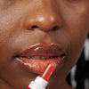 Novexpert HYALURONIC Lip'Up Huulivoide; lähikuva henkilön kasvoista, huulet kiiltävät sovellettaessa kirkasta huulikiiltoa pienellä tuubiapplikaattorilla; ihonväri korostuu kuvassa; tausta epätarkka, tuote ja levitysliike kuvan keskiössä.
