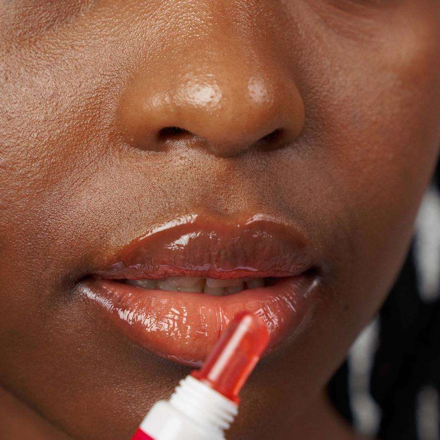 Novexpert HYALURONIC Lip'Up Huulivoide; lähikuva henkilön kasvoista, huulet kiiltävät sovellettaessa kirkasta huulikiiltoa pienellä tuubiapplikaattorilla; ihonväri korostuu kuvassa; tausta epätarkka, tuote ja levitysliike kuvan keskiössä.