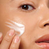 Novexpert MAGNESIUM Hydro-Biotic Velvety Cream; lähikuva henkilöstä levittämässä valkoista voidetta poskelleen sormin, näkyvissä osa silmästä ja huulista, iho sileän ja säteilevän näköinen; ei näkyvää pakkausta tai brändäystä; neutraali tausta.