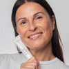 Novexpert OMEGA Rich Protective Cream; hymyilevä henkilö suora ruskea tukka pitelee kosteuttavaa ihonhoitovoidetuubia lähellä kasvojaan neutraalia taustaa vasten; tuubi valkoinen ja sylinterinmuotoinen mattapinnalla ja kierrekorkilla; etuetiketissä vähäeleistä groteskitekstiä; ei muita esineitä näkyvissä, keskikokoinen lähikuva.