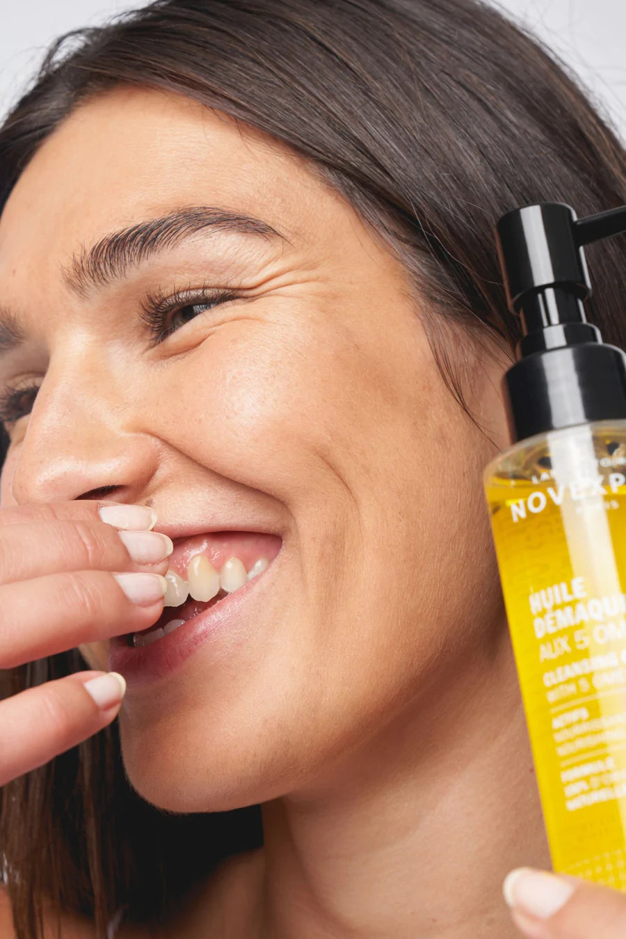 Novexpert OMEGA Cleansing Oil with 5 Omegas; hymyilevä pitkätukkainen ruskeahiuksinen henkilö pitelee pulloa lähellä kasvojaan, toinen käsi koskettaa suuta; pullo sylinterinmuotoinen muovia kiiltäväpintaisena mustalla korkilla, etiketissä valkoinen versaaliteksti osin lihavoituna; vaaleanharmaa tausta, keskisuuri lähikuva.