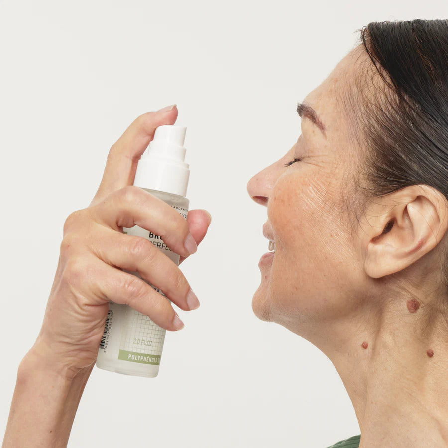 Novexpert POLYPHENOLS Perfect Glow Mist; iäkkäämpi nainen hymyilee silmät suljettuina nauttien raikastavasta ihonhoitosuihkeesta valkoisesta pullosta valkoisella taustalla; pullo muovinen, lieriön muotoinen ja mattapintainen ilman näkyvää tekstiä; kuvattu vyötäröstä ylöspäin.