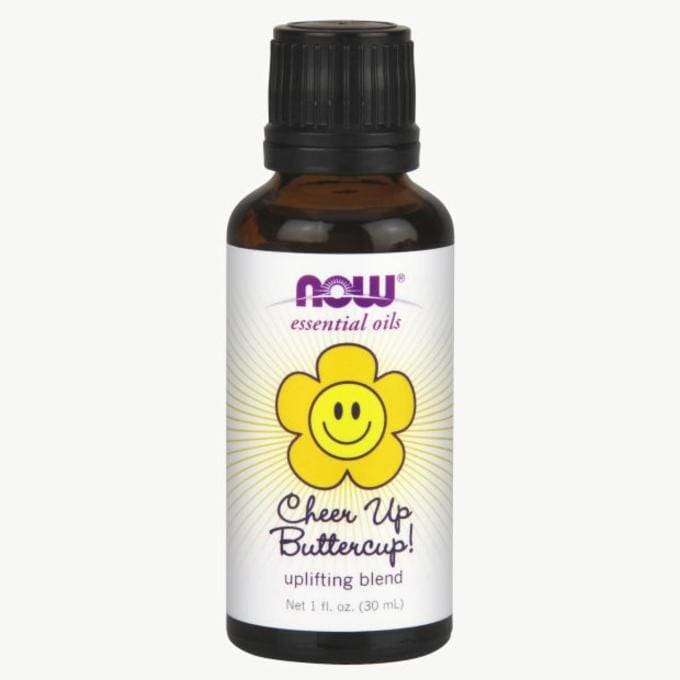 NOW Essential Oils Cheer Up Buttercup! -eteerisen öljyn seos, 1 oz; tummanruskea, lieriömäinen lasipullo, musta uritettu korkki; etiketissä hymyilevä keltainen kukka ja teksti "uplifting blend"; etiketin pohjaväri pääosin valkoinen, painettuja graafisia elementtejä ja groteskitekstiä; vaalea tausta, yksinkertainen studiokuva, ei ihmisiä.