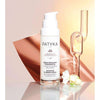 Patyka Advanced Plumping Serum -pullo pystyssä mattapersikanvärisellä alustalla; pullo sylinterimäinen, kirkasta tai huurrettua lasia, valkoinen korkki; etiketissä "Patyka Advanced Plumping Serum" yksinkertaisella mustalla fontilla; lähellä kirkkaita lasikoriste-esineitä ja pieniä valkoisia kukkia; tausta jaettu persikan- ja beigensävyihin; ei ihmisiä näkyvissä.