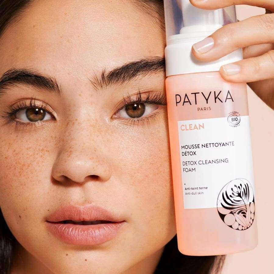 Pisamainen nainen pitelee sylinterimäistä valkoista muovipulloa, jossa lukee "Patyka Paris Clean Detox Cleansing Foam" mustalla tekstausvapaalla tekstillä, kasvojensa vieressä; pullo mattapintainen pumppuannostelijalla; kuva lähikuva yksinkertaista taustaa vasten; nainen esittelee ihonpuhdistustuotteen kasvojensa vieressä.