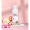 Patyka Detox Cleansing Foam -pullo, sylinterimäinen valkoinen muovirunko ja läpinäkyvä korkki, pystyssä suorakulmaisella kiiltävällä valkoisella tarjottimella; etiketti pääosin valkoinen mustalla tekstausvapaalla tekstillä, tuotenimi ja brändi näkyvissä; vaaleanpunaisia ja persikanvärisiä kukkia tarjottimen ympärillä; tausta yhtenäisen pinkki; ei ihmisiä tai käsiä näkyvissä; kuva suora edestä, koko tuote näkyvissä.