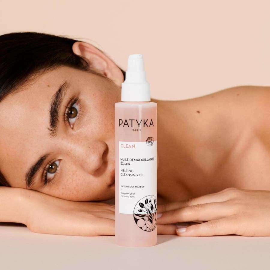 Pisamainen nainen nojaa päätään käsivarteensa Patyka Paris Clean Melting Cleansing Oil -pullon vieressä, joka esillä etualalla beigeä taustaa vasten; pullo kirkas ja sylinterimäinen pumppukorkilla, sisältää vaaleankeltaista nestettä; etiketissä tuote- ja brändinimi tekstausvapaalla fontilla; kuva pehmeästi valaistu ja tiiviisti rajattu henkilön ja tuotteen ympärille.